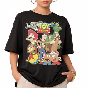 May include: Black t-shirt featuring a colorful graphic of characters from the animated film "Toy Story." The design includes Woody, Buzz Lightyear, Jessie, Rex, and other characters, with the words "Toy Story" in a vibrant font.