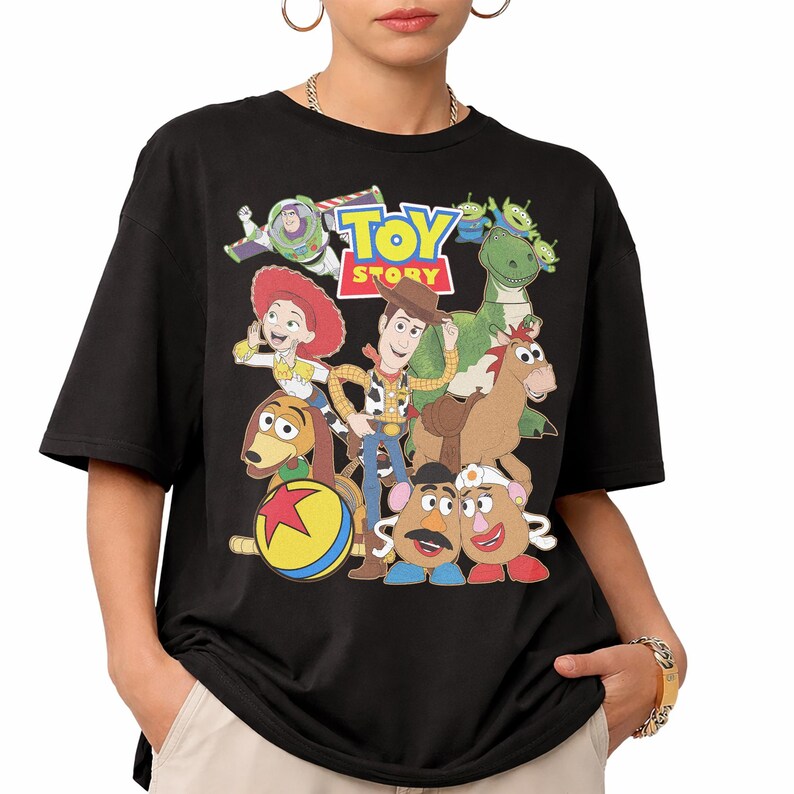 May include: Black t-shirt featuring a colorful graphic of characters from the animated film "Toy Story." The design includes Woody, Buzz Lightyear, Jessie, Rex, and other characters, with the words "Toy Story" in a vibrant font.