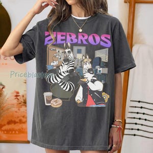 Pu&ograve; includere: T-shirt grigio scuro con un'illustrazione a cartoni animati di due zebre in un ufficio. La parola "ZEBROS" &egrave; stampata in alto in un carattere sfumato viola e rosa. Una zebra indossa una giacca, l'altra un abito.