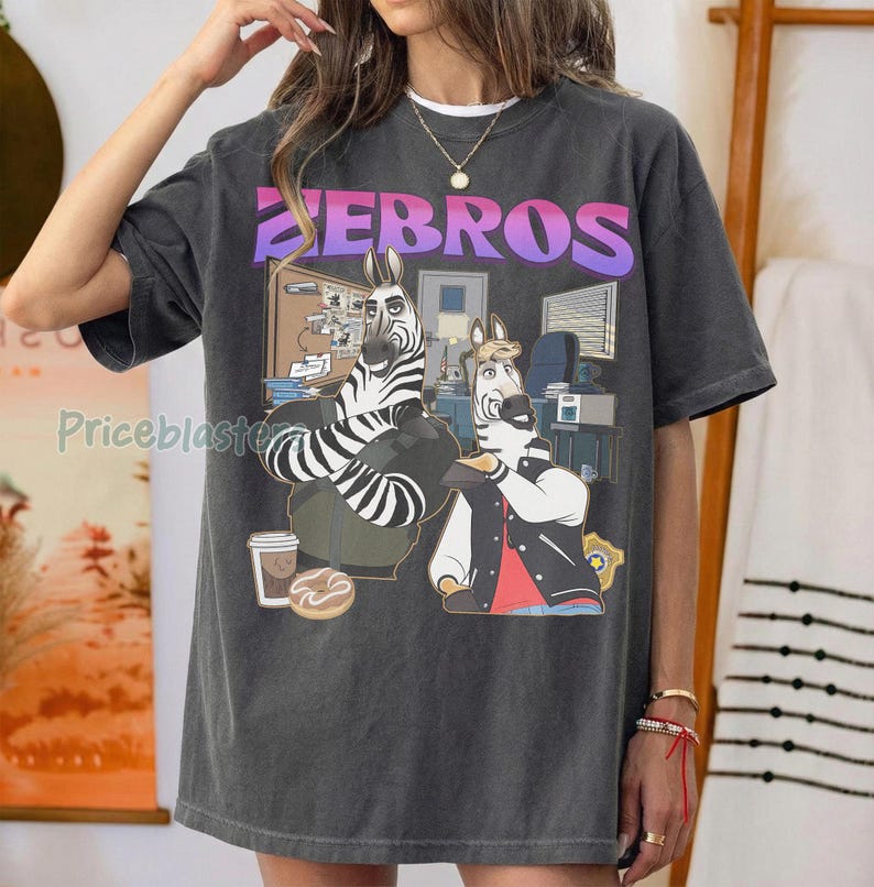 Pu&ograve; includere: T-shirt grigio scuro con un'illustrazione a cartoni animati di due zebre in un ufficio. La parola "ZEBROS" &egrave; stampata in alto in un carattere sfumato viola e rosa. Una zebra indossa una giacca, l'altra un abito.