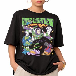 May include: Black t-shirt featuring a graphic of Buzz Lightyear in a space scene with the text "Buzz Lightyear Lazer Blast" in green and white. The design includes cartoon characters, planets, and stars.