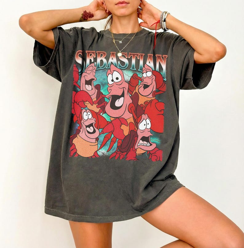Pu&ograve; includere: T-shirt grigio scuro con una grafica del personaggio Sebastian de La Sirenetta. L'immagine mostra Sebastian e altri granchi rossi con il nome "Sebastian" in un font colorato. La maglietta ha maniche corte e una vestibilit&agrave; ampia.