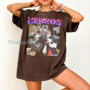 Pu&ograve; includere: T-shirt marrone con la scritta "ZEBROS" in lettere viola e bianche. La grafica presenta due zebre dei cartoni animati in un ambiente d'ufficio. Una zebra indossa una giacca e l'altra &egrave; seduta su una sedia.