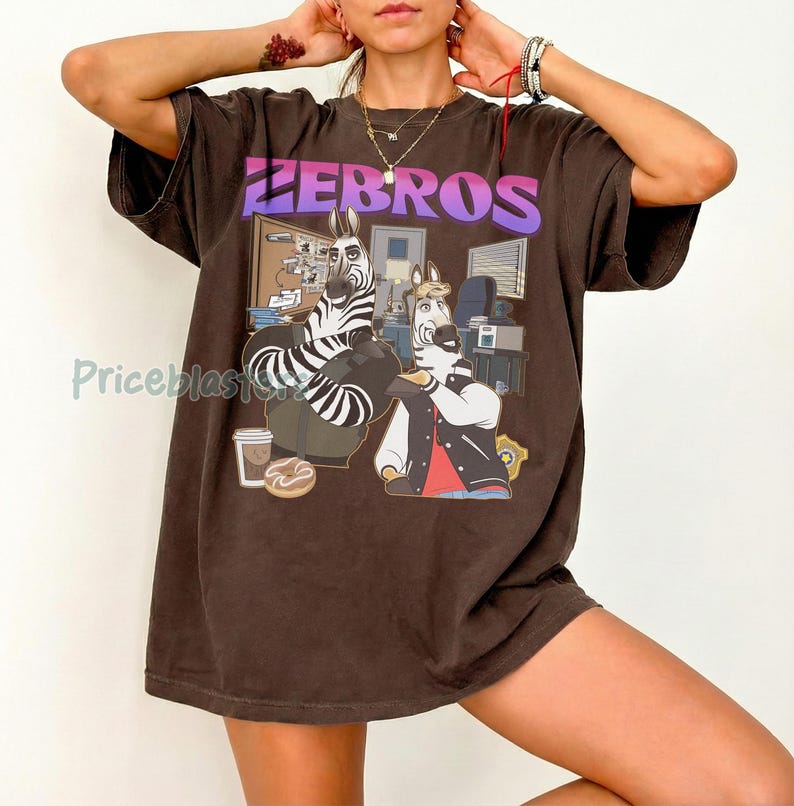 Pu&ograve; includere: T-shirt marrone con la scritta "ZEBROS" in lettere viola e bianche. La grafica presenta due zebre dei cartoni animati in un ambiente d'ufficio. Una zebra indossa una giacca e l'altra &egrave; seduta su una sedia.