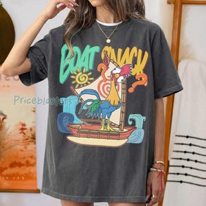 Puede incluir: Camiseta gris oscuro con un gr&aacute;fico colorido de un gallo navegando en un barco, con las palabras "BOAT SNACK" en una fuente l&uacute;dica. El dise&ntilde;o incluye un sol, olas y signos de interrogaci&oacute;n.