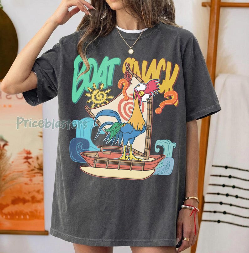 Puede incluir: Camiseta gris oscuro con un gr&aacute;fico colorido de un gallo navegando en un barco, con las palabras "BOAT SNACK" en una fuente l&uacute;dica. El dise&ntilde;o incluye un sol, olas y signos de interrogaci&oacute;n.