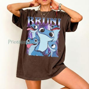 May include: A brown t-shirt featuring the word "BRUNI" in a purple and blue font. The shirt displays a graphic of several blue salamander-like creatures with large eyes and tongues. The design is set against a purple lightning background.