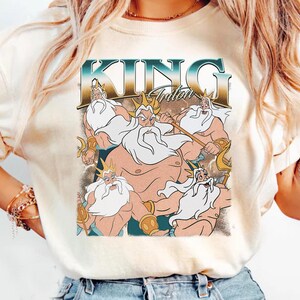 May include: Cream-colored t-shirt featuring a graphic of King Triton from The Little Mermaid. The design includes the text "KING Triton" in a teal and gold font, with illustrations of Triton holding a trident.