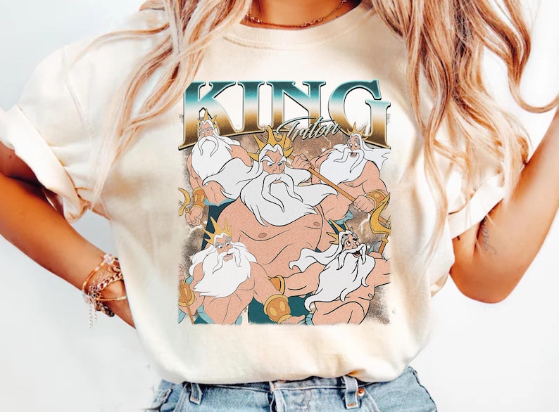 May include: Cream-colored t-shirt featuring a graphic of King Triton from The Little Mermaid. The design includes the text "KING Triton" in a teal and gold font, with illustrations of Triton holding a trident.