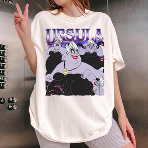 May include: Off-white t-shirt featuring a graphic of Ursula from The Little Mermaid. The graphic includes the name "URSULA" in purple and images of Ursula in various poses. The shirt is a relaxed fit.