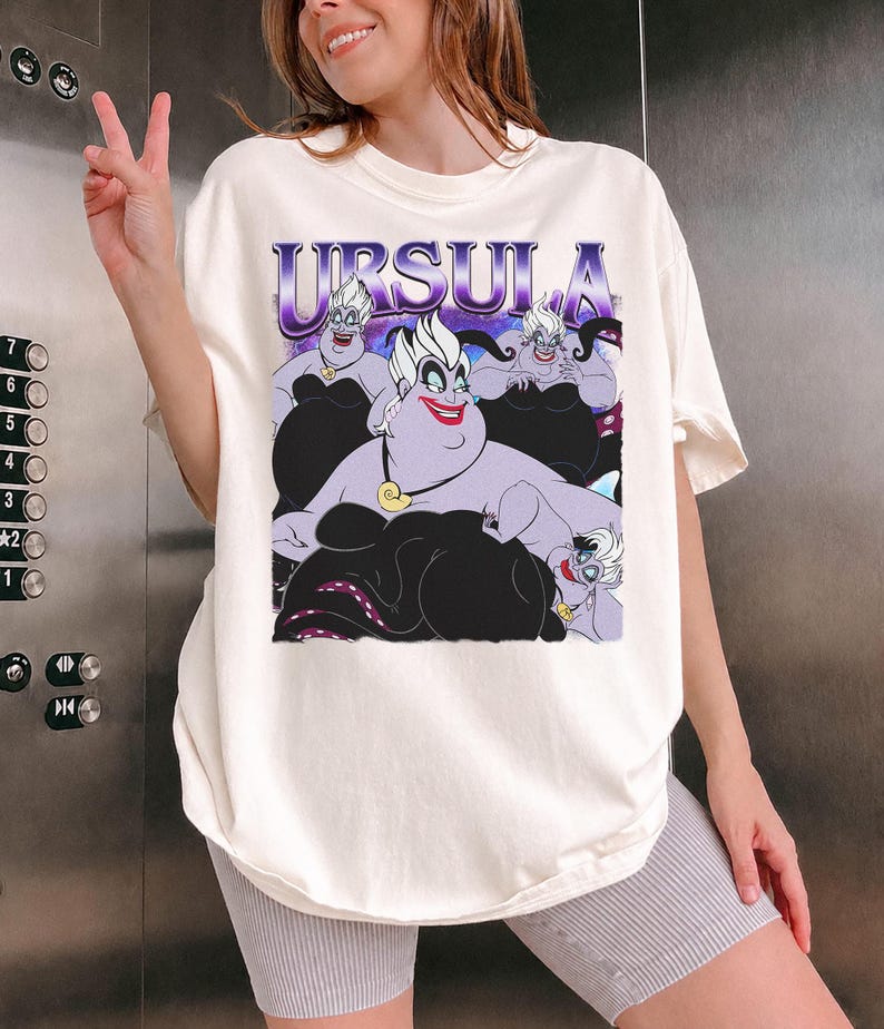 May include: Off-white t-shirt featuring a graphic of Ursula from The Little Mermaid. The graphic includes the name "URSULA" in purple and images of Ursula in various poses. The shirt is a relaxed fit.