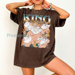 May include: Brown t-shirt featuring a graphic of King Triton from The Little Mermaid. The design includes the text "KING Triton" in a blue and gold font, with an illustration of the character and other figures.