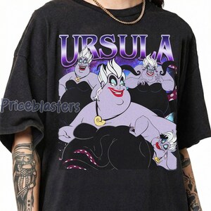 May include: Dark gray t-shirt featuring a graphic of Ursula from The Little Mermaid. The design includes multiple images of Ursula with the word "URSULA" in a purple and white font. The shirt has a crew neck and short sleeves.