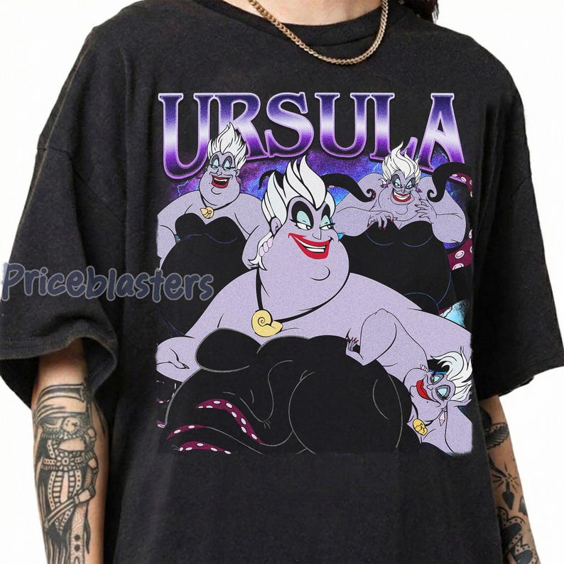 May include: Dark gray t-shirt featuring a graphic of Ursula from The Little Mermaid. The design includes multiple images of Ursula with the word "URSULA" in a purple and white font. The shirt has a crew neck and short sleeves.