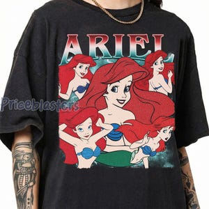 Pu&ograve; includere: T-shirt grigio scuro con la scritta "ARIEL" in rosso e blu. La maglietta presenta diverse illustrazioni di una sirena dai capelli rossi con un reggiseno a conchiglia blu. Il design ha un aspetto vintage e usurato.