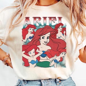 Pu&ograve; includere: T-shirt color crema con una grafica di Ariel de La Sirenetta. Il design include pi&ugrave; immagini di Ariel con capelli rossi e un reggiseno a conchiglia blu, con il nome "ARIEL" in un font retr&ograve;.