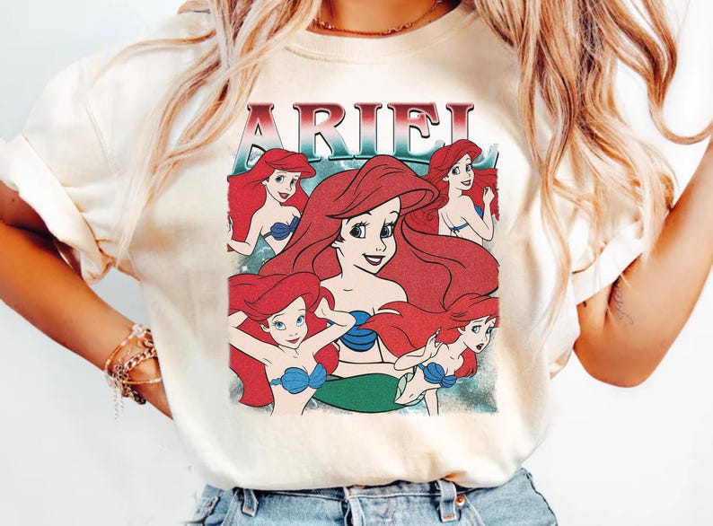 Pu&ograve; includere: T-shirt color crema con una grafica di Ariel de La Sirenetta. Il design include pi&ugrave; immagini di Ariel con capelli rossi e un reggiseno a conchiglia blu, con il nome "ARIEL" in un font retr&ograve;.