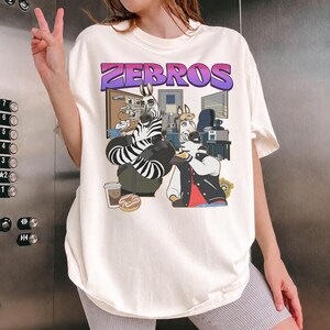 Pu&ograve; includere: T-shirt color crema con una grafica a cartoni animati di zebre in un ambiente d'ufficio, con la scritta "ZEBROS" in viola e rosa. La maglietta &egrave; indossata da una persona che fa il segno della pace.