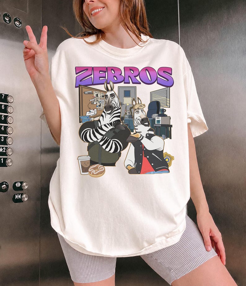 Pu&ograve; includere: T-shirt color crema con una grafica a cartoni animati di zebre in un ambiente d'ufficio, con la scritta "ZEBROS" in viola e rosa. La maglietta &egrave; indossata da una persona che fa il segno della pace.