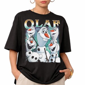 Puede incluir: Camiseta negra con un gr&aacute;fico de Olaf de Frozen. El dise&ntilde;o presenta varios personajes de Olaf con narices de zanahoria y caras sonrientes. La palabra "OLAF" est&aacute; impresa en la parte superior en una fuente estilizada.