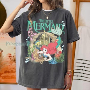 Może przedstawiać: Ciemnoszara koszulka z kolorową grafiką z Małej Syrenki Disneya. Projekt zawiera Ariel, Florka, Sebastiana i zamek. Tekst "The Little Mermaid" jest w kolorze zielonym.