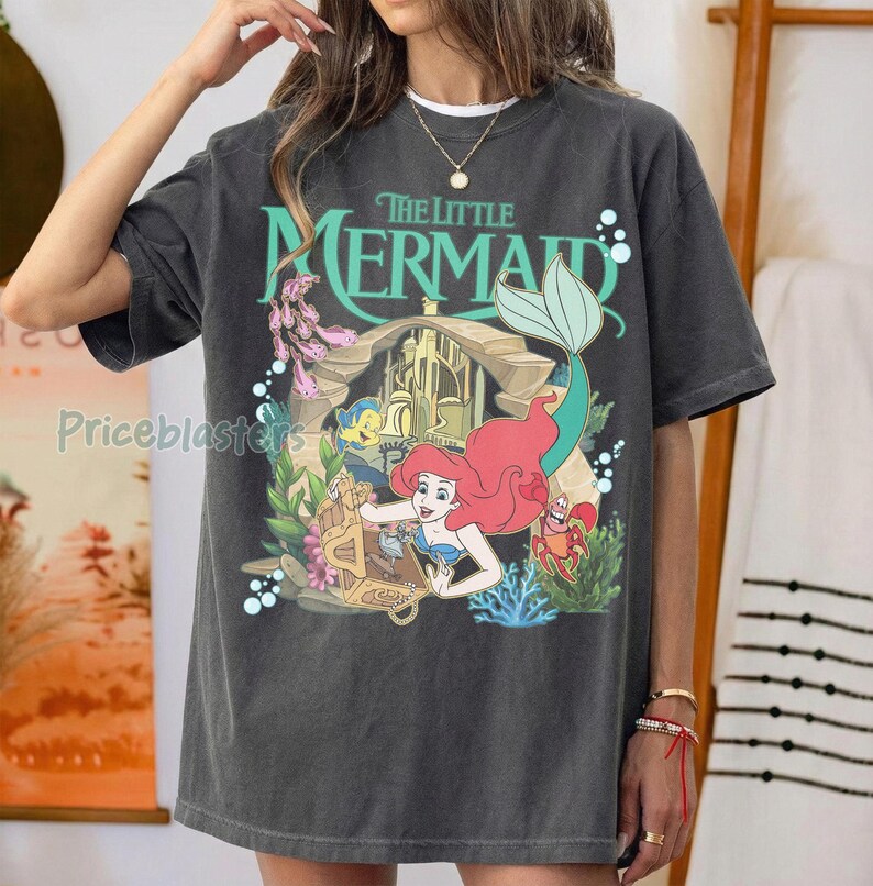 Może przedstawiać: Ciemnoszara koszulka z kolorową grafiką z Małej Syrenki Disneya. Projekt zawiera Ariel, Florka, Sebastiana i zamek. Tekst "The Little Mermaid" jest w kolorze zielonym.