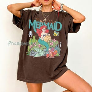May include: Brown t-shirt featuring a graphic of Ariel from Disney's The Little Mermaid. The design includes Ariel with red hair, a turquoise tail, and the text "The Little Mermaid" in turquoise and orange.