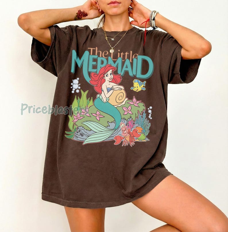 May include: Brown t-shirt featuring a graphic of Ariel from Disney's The Little Mermaid. The design includes Ariel with red hair, a turquoise tail, and the text "The Little Mermaid" in turquoise and orange.