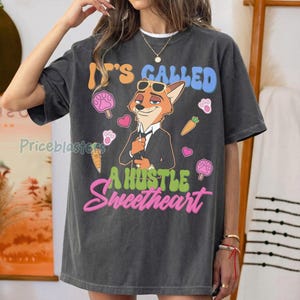 May include: Dark gray t-shirt featuring a cartoon fox in a suit with sunglasses. The shirt has the text "It's Called A Hustle Sweetheart" in blue, green, and pink. Additional graphics include paw prints, hearts, and carrots.