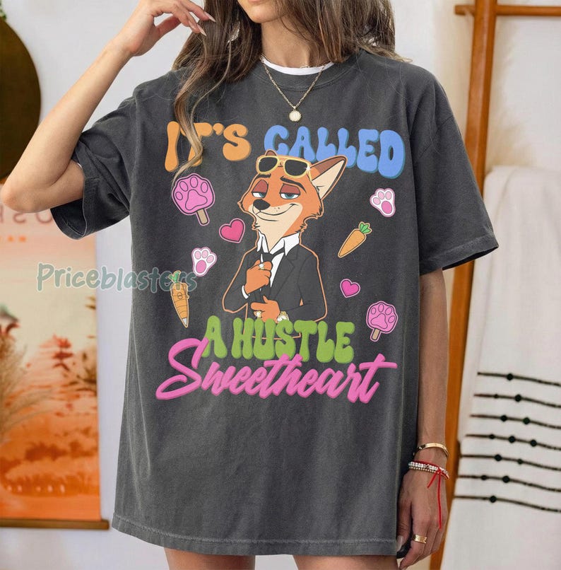 May include: Dark gray t-shirt featuring a cartoon fox in a suit with sunglasses. The shirt has the text "It's Called A Hustle Sweetheart" in blue, green, and pink. Additional graphics include paw prints, hearts, and carrots.