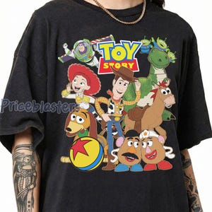 May include: Dark gray t-shirt featuring a colorful graphic of characters from the animated film "Toy Story." The design includes Woody, Buzz Lightyear, Jessie, Rex, and other characters, with the "Toy Story" logo at the top.