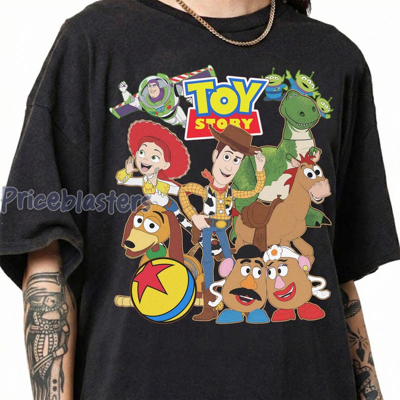 May include: Dark gray t-shirt featuring a colorful graphic of characters from the animated film "Toy Story." The design includes Woody, Buzz Lightyear, Jessie, Rex, and other characters, with the "Toy Story" logo at the top.