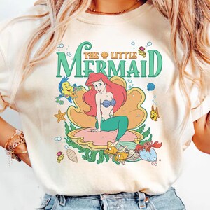 May include: Cream-coloured t-shirt with a colourful graphic of Ariel from The Little Mermaid. The design features Ariel in a seashell, surrounded by sea creatures and the words "The Little Mermaid" in green and gold.