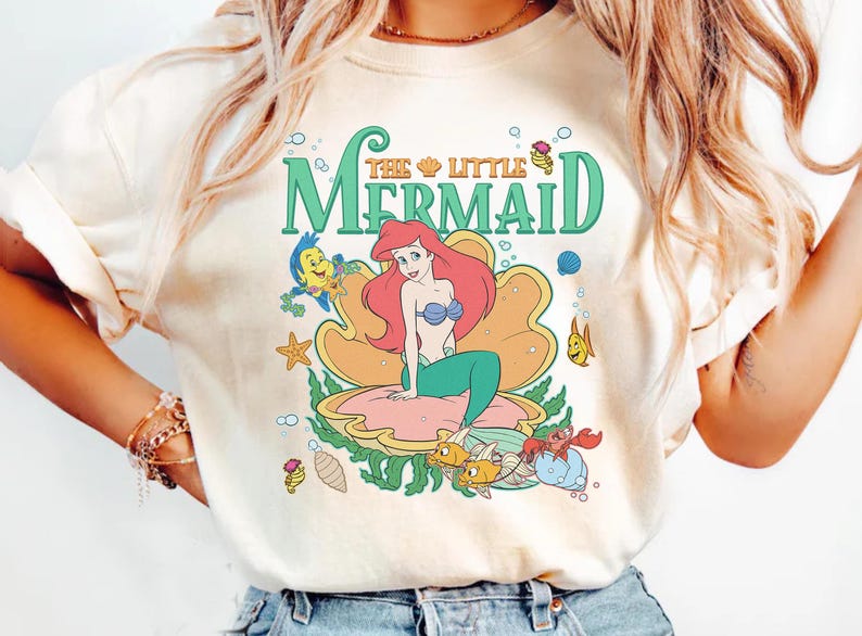 May include: Cream-coloured t-shirt with a colourful graphic of Ariel from The Little Mermaid. The design features Ariel in a seashell, surrounded by sea creatures and the words "The Little Mermaid" in green and gold.