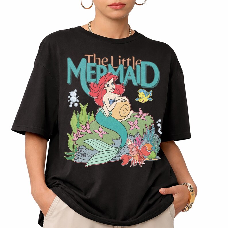 May include: Black t-shirt featuring a colorful graphic of Ariel from The Little Mermaid. The design includes Ariel, Flounder, and Sebastian, with the words "The Little Mermaid" above. The shirt has short sleeves and a crew neck.