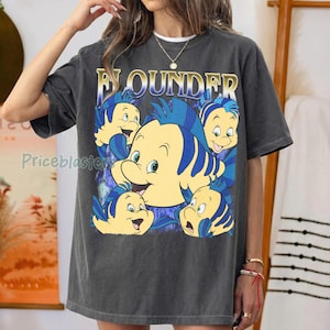 May include: Dark gray t-shirt featuring the word "Flounder" in gold lettering above a cartoon image of the character Flounder from The Little Mermaid. The graphic includes multiple yellow and blue fish.