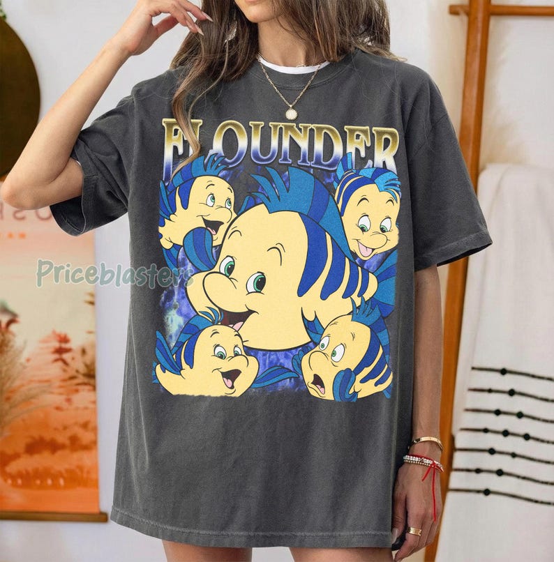 May include: Dark gray t-shirt featuring the word "Flounder" in gold lettering above a cartoon image of the character Flounder from The Little Mermaid. The graphic includes multiple yellow and blue fish.