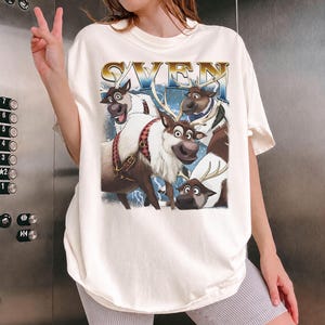 May include: Cream-colored t-shirt featuring a graphic of the character Sven from the movie Frozen. The word "SVEN" is printed in gold letters above the image of the reindeer. The image includes other reindeer characters.
