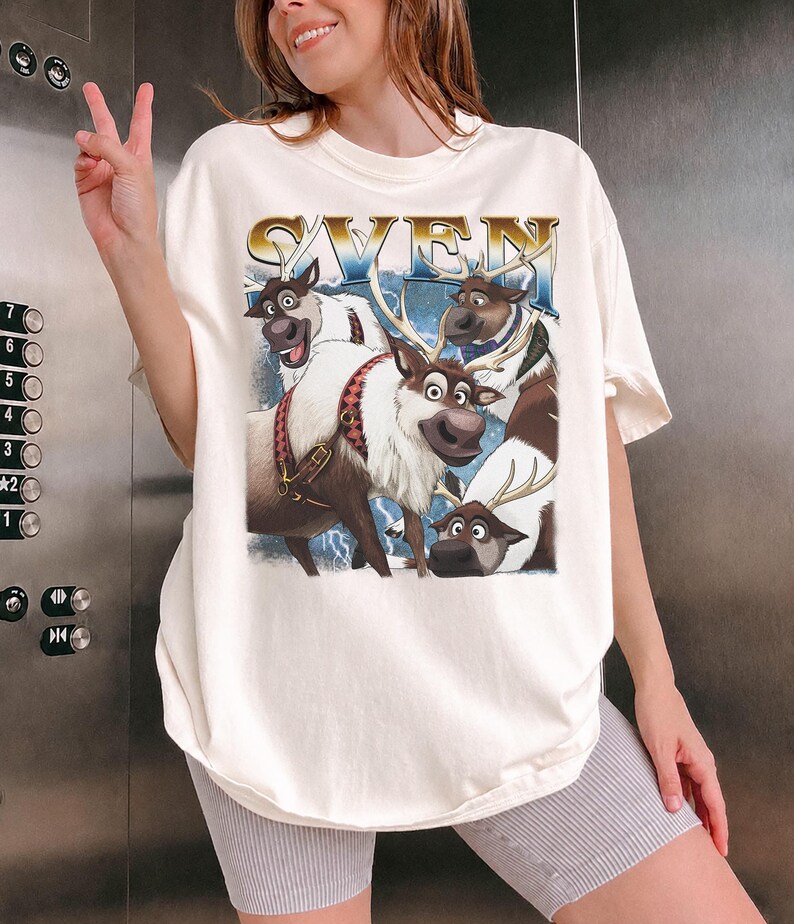 May include: Cream-colored t-shirt featuring a graphic of the character Sven from the movie Frozen. The word "SVEN" is printed in gold letters above the image of the reindeer. The image includes other reindeer characters.