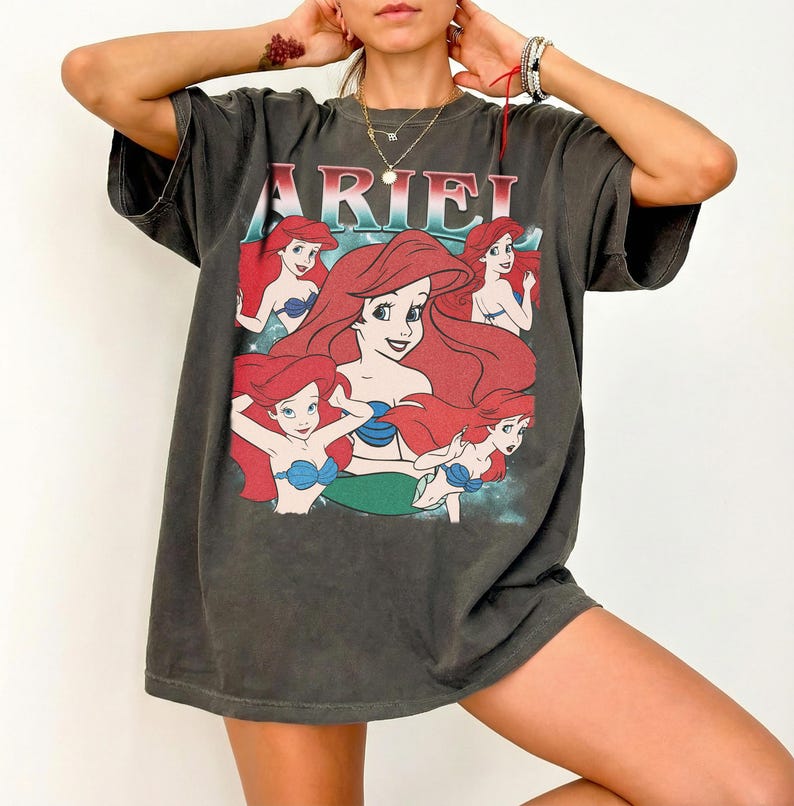 Pu&ograve; includere: T-shirt grigio scuro con la scritta "ARIEL" in rosso e una grafica di Ariel de La Sirenetta. La grafica mostra Ariel con i capelli rossi e un top blu. La maglietta ha maniche corte e girocollo.