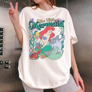 May include: Cream-colored t-shirt featuring a colorful graphic from "The Little Mermaid." The design includes Ariel, a dolphin, and other characters from the movie, with the title in a teal font. The shirt is a relaxed fit.