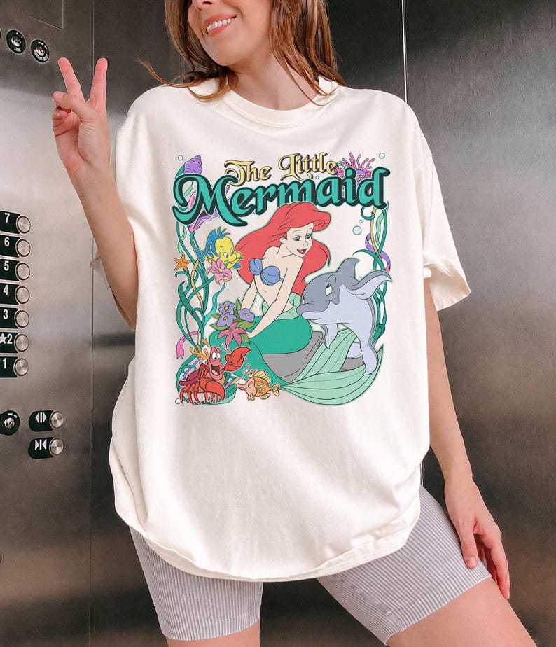 May include: Cream-colored t-shirt featuring a colorful graphic from "The Little Mermaid." The design includes Ariel, a dolphin, and other characters from the movie, with the title in a teal font. The shirt is a relaxed fit.