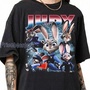 Op de afbeelding: Donkergrijze t-shirt met een kleurrijke afbeelding van Judy Hopps en andere personages uit Zootopia. Het woord "JUDY" staat in grote, gestileerde letters boven de personages. Het shirt heeft een ronde hals en korte mouwen.