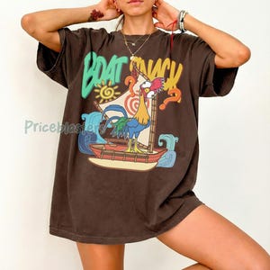 Puede incluir: Camiseta marr&oacute;n con un gr&aacute;fico colorido de un gallo navegando en un barco, con las palabras "BOAT SNACK" en una fuente juguetona. El dise&ntilde;o incluye un sol, olas y signos de interrogaci&oacute;n. La camiseta tiene mangas cortas.