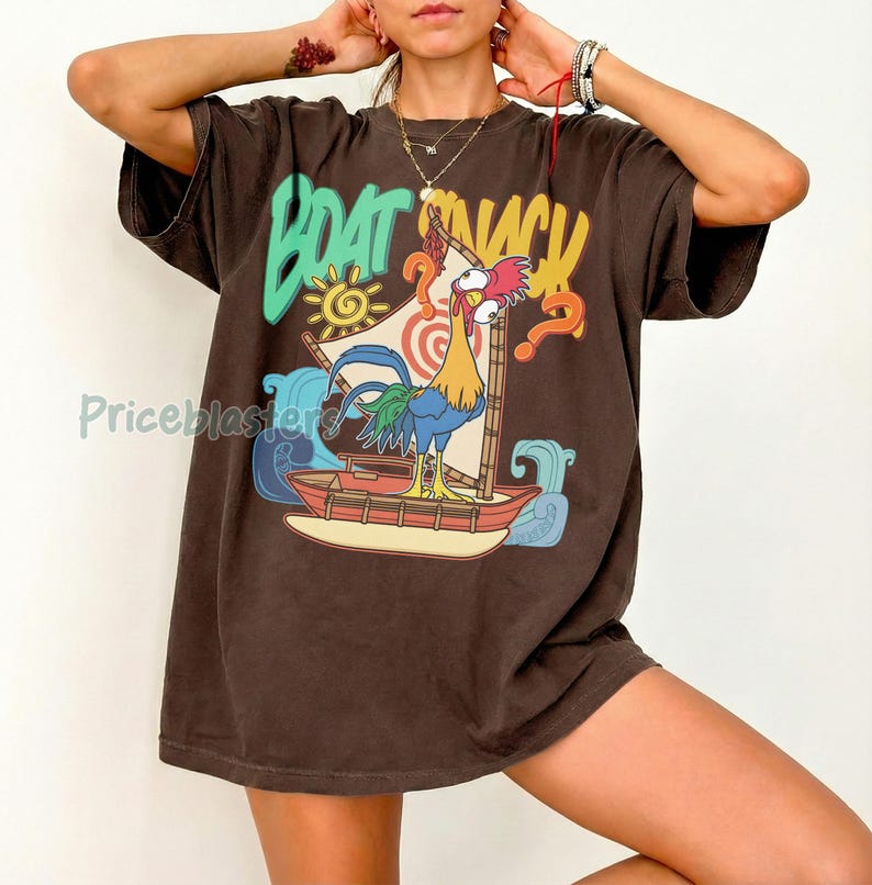 Puede incluir: Camiseta marr&oacute;n con un gr&aacute;fico colorido de un gallo navegando en un barco, con las palabras "BOAT SNACK" en una fuente juguetona. El dise&ntilde;o incluye un sol, olas y signos de interrogaci&oacute;n. La camiseta tiene mangas cortas.