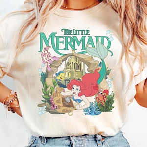 Może przedstawiać: Kremowa koszulka z kolorową grafiką z Małej Syrenki Disneya. Projekt zawiera Ariel, Florka, Sebastiana i napis "The Little Mermaid" w turkusowym kolorze. Grafika otoczona jest elementami podwodnymi.