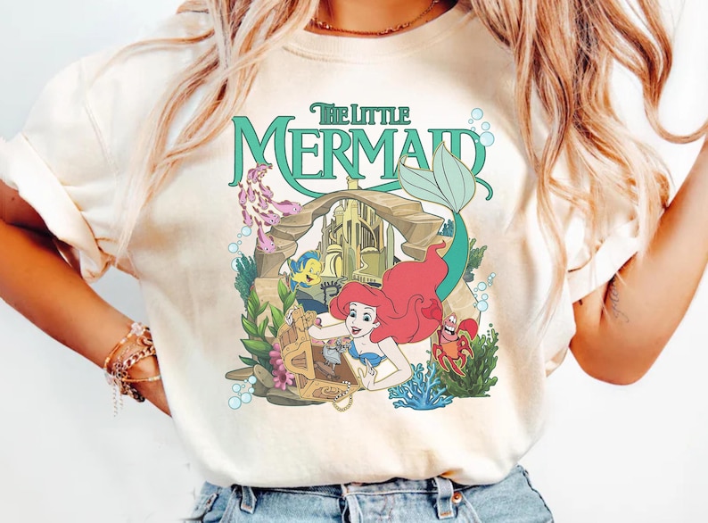 Może przedstawiać: Kremowa koszulka z kolorową grafiką z Małej Syrenki Disneya. Projekt zawiera Ariel, Florka, Sebastiana i napis "The Little Mermaid" w turkusowym kolorze. Grafika otoczona jest elementami podwodnymi.