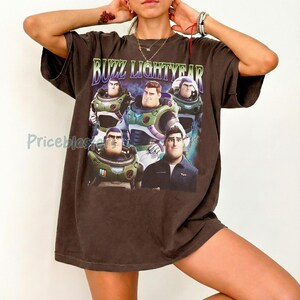 May include: Dark brown Buzz Lightyear graphic t-shirt. The shirt features a graphic of Buzz Lightyear and the text "Buzz Lightyear" in green and purple. The shirt is oversized and has short sleeves.