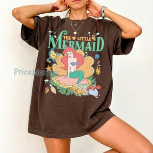 May include: Brown t-shirt with a graphic of Ariel from The Little Mermaid. The design shows Ariel on a seashell, surrounded by sea creatures, with the words "The Little Mermaid" in green and gold.