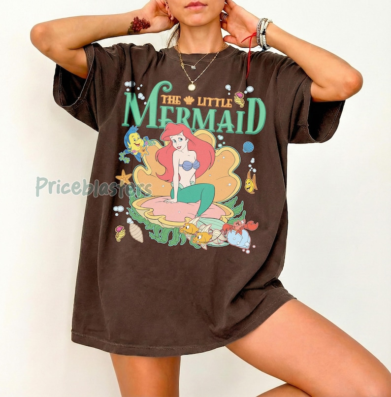 May include: Brown t-shirt with a graphic of Ariel from The Little Mermaid. The design shows Ariel on a seashell, surrounded by sea creatures, with the words "The Little Mermaid" in green and gold.